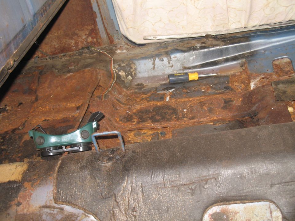 Heavily rusted interior floor pan of a vehicle with a green seatbelt buckle and screwdriver.