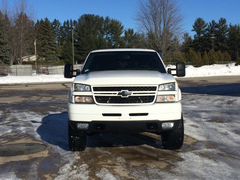 2006 Chevrolet Silverado 2500 - Front view of a white Chevrolet Silverado 2500 pickup truck parked on a wet, icy surface with snow in the background. The truck has large t…