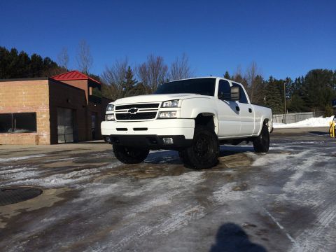 2006 Chevrolet Silverado 2500 - A lifted white Chevrolet Silverado 2500 pickup truck is parked on a wet, icy lot in front of a brick building with a red roof. The truck is…
