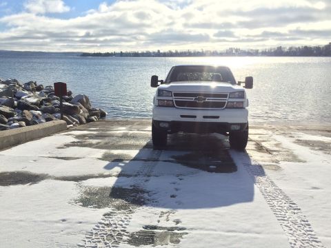 2006 Chevrolet Silverado 2500 - A white Chevrolet pickup truck is parked on a snowy boat ramp facing the water. The sun is shining brightly over the lake.