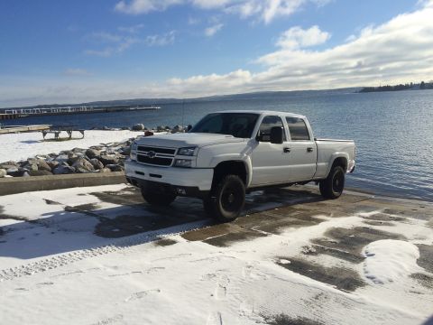 2006 Chevrolet Silverado 2500 - A white Chevrolet Silverado pickup truck is parked on a wooden dock next to a body of water on a partly cloudy day. The ground is covered i…