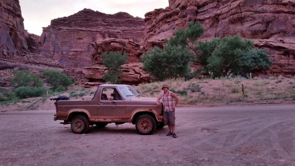 Brown 1987 Ford Bronco II parked on a dirt road with a man leaning on the fender. Red rock cliffs and green trees in the background