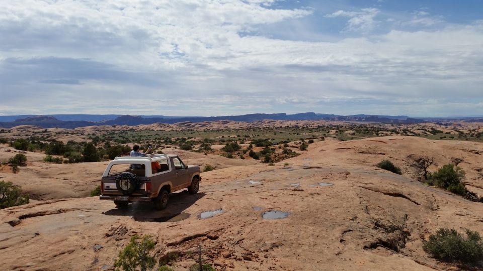 1980s Ford Bronco with spare tire on rear door, driving on rocky desert terrain under a cloudy sky