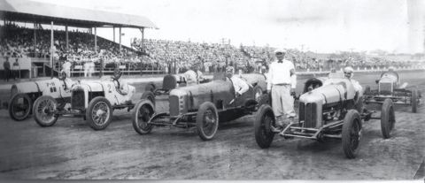 1909 Ford Model T - Several vintage race cars are parked on a dirt track in front of a grandstand filled with spectators.