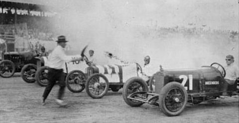 1909 Ford Model T - Multiple race cars are lined up on a track, with a man in the foreground and spectators in the background.