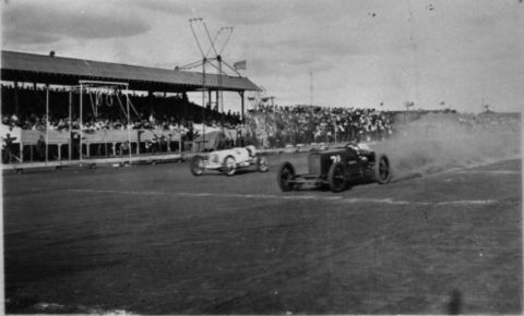 1909 Ford Model T - A pair of early 20th-century race cars speed around a track before a large crowd.