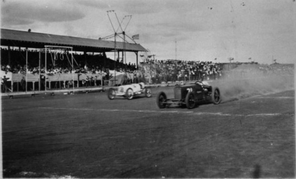 Two open-wheel race cars compete on a dirt track, one kicking up dust
