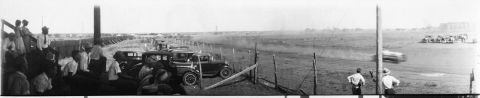 1909 Ford Model T - Spectators watch cars compete on a dusty track, with several vehicles visible in the background and foreground.