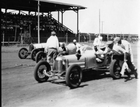 1909 Ford Model T - Race cars prepare for a competition at a track, with a crowd watching from the stands.