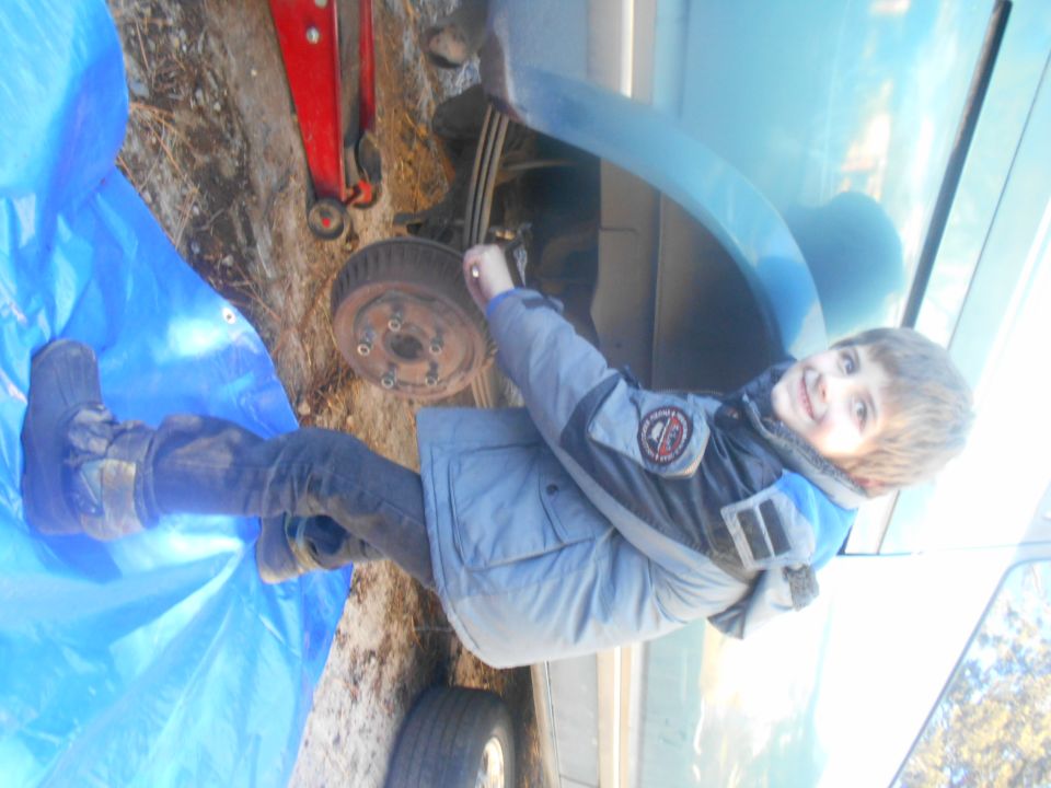 Child in jacket and jeans working on a car's rear brake drum, which is exposed