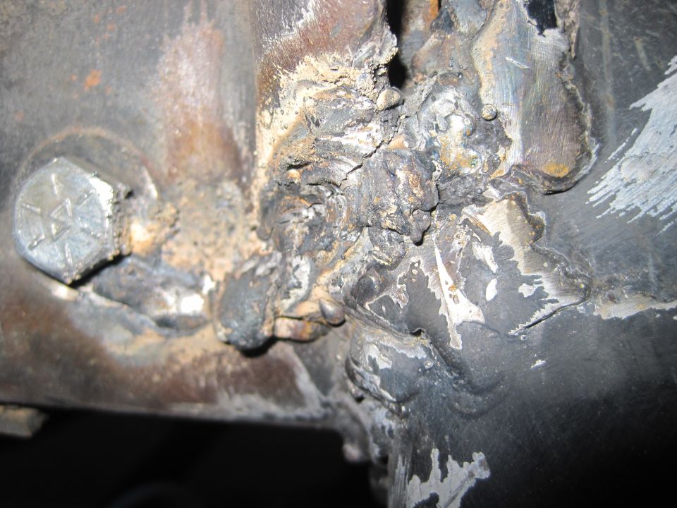 Close-up of a rusty, welded metal joint with a hex bolt. The metal is corroded and shows signs of repair