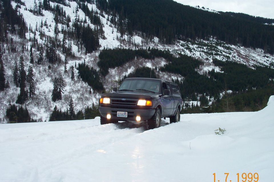 Dark Ford pickup with canopy driving through deep snow on a mountain road, lights on
