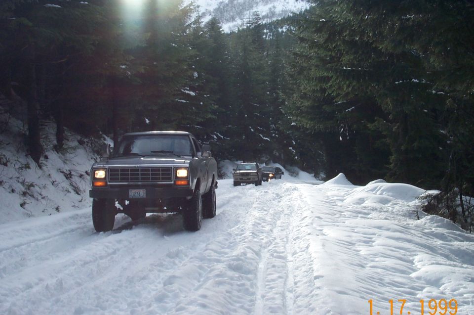 Lifted gray pickup with headlights on leading two vehicles down a snowy tree-lined mountain road