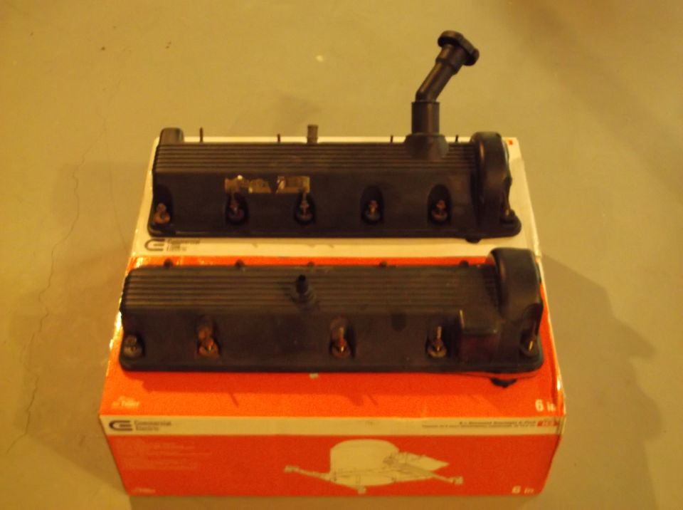 Two black engine valve covers stacked on an orange box. One has a black hose fitting