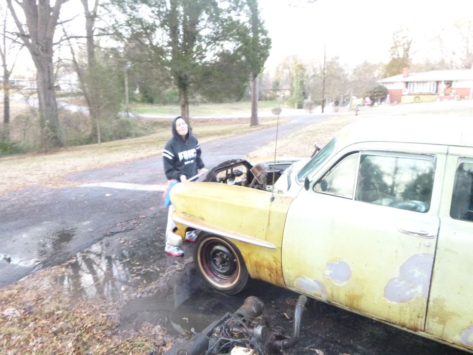 Person standing next to a yellow 1953 Plymouth Belvedere with the hood open and engine removed