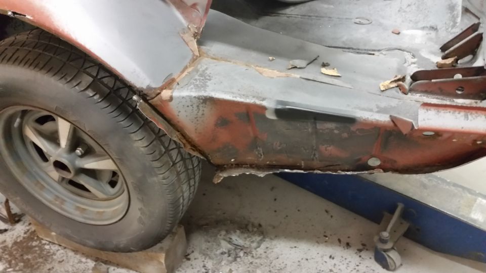 Red car body panel with rust and damage, showing a tire and wheel. The vehicle is on a lift