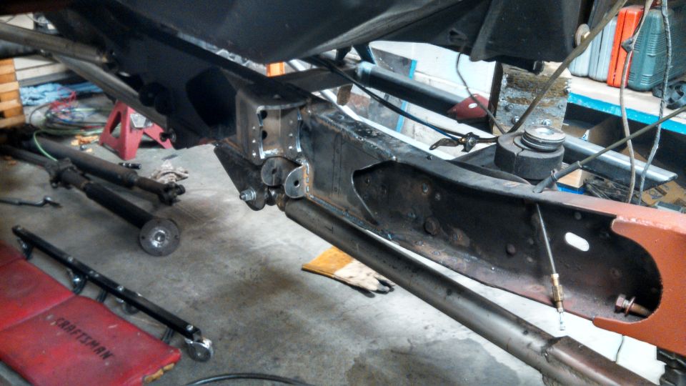 Close-up of a truck frame with driveshafts laid out nearby. A red Craftsman creeper is visible