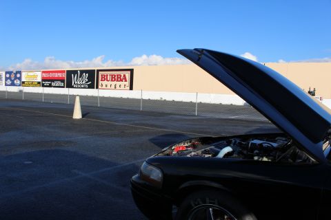 2003 Ford Crown Victoria - black car with its hood raised sits on a paved drag strip, with advertising banners including Bubba Burger on the wall behind.
