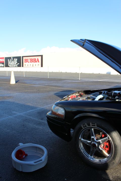 2003 Ford Crown Victoria - Front quarter of a black car with the hood raised and a chrome five-spoke wheel, parked on a drag strip near banners.