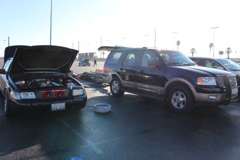 2003 Ford Crown Victoria - black sedan with its hood raised parked beside a black-and-tan Ford Expedition SUV on a sunlit asphalt lot.