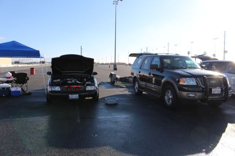 2003 Ford Crown Victoria - black sedan with raised hood sits beside a black Ford Expedition next to a blue pop-up canopy and folding chairs in a lot.