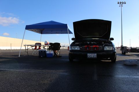 2003 Ford Crown Victoria - front of a black sedan with its hood up showing twin red components, plate reading EVIL VIC, next to a blue canopy.