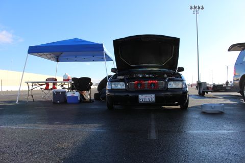 2003 Ford Crown Victoria - black sedan with its hood up and EVIL VIC plate parked beside a blue pop-up tent and folding chairs in a paved lot.
