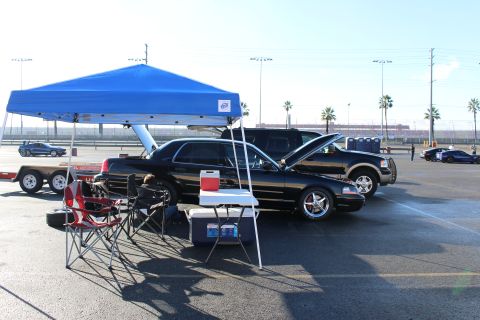 2003 Ford Crown Victoria - black sedan parked under a blue canopy with chairs and a cooler, an SUV and palm trees in the background of a lot.