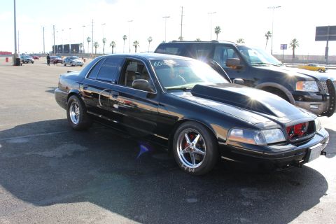 2003 Ford Crown Victoria - lowered black sedan with chrome five-spoke wheels parked beside a dark SUV in a palm-lined asphalt lot.