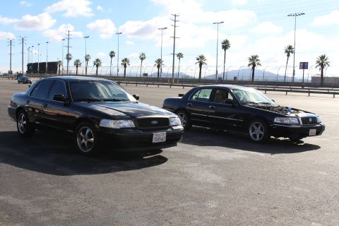 2003 Ford Crown Victoria - Two black Ford sedans parked at angles in an open palm-lined parking lot under a partly cloudy blue sky.