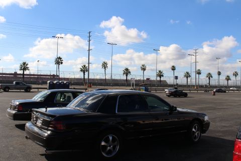 2003 Ford Crown Victoria - Rear view of a black sedan parked in a large lot with palm trees, stadium lights and a cloudy blue sky behind.