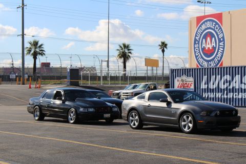 2003 Ford Crown Victoria - black sedan and a gray Ford Mustang parked beside shipping containers with AAA and Lucas Oil signage in a lot.