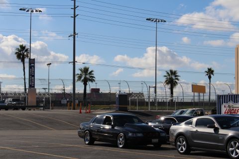 2003 Ford Crown Victoria - black sedan and a gray Mustang parked at the edge of a lot with a racetrack grandstand and fencing in the background.