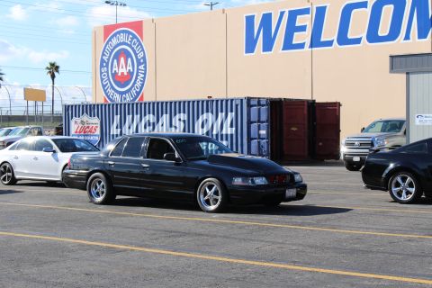 2003 Ford Crown Victoria - lowered black sedan with chrome wheels parked among other cars in front of a building with AAA emblem and WELCOME sign.
