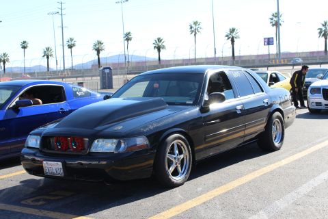 2003 Ford Crown Victoria - lowered black Ford sedan with chrome five-spoke wheels and red components visible behind its grille in a sunny lot.