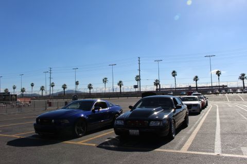 2003 Ford Crown Victoria - dark blue Ford Mustang sits beside a black full-size sedan in a sunlit parking lot with palm trees and a racetrack in the background.