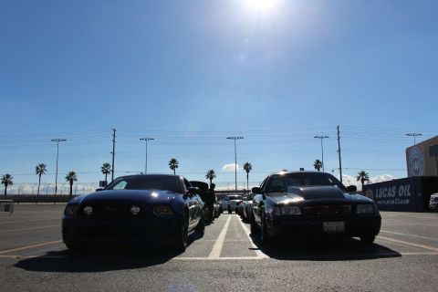 2003 Ford Crown Victoria - blue Ford Mustang and a black sedan face the camera in a bright parking lot, sun flaring overhead with palm trees and signage behind.