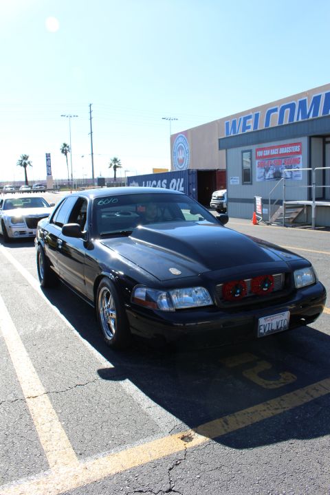 2003 Ford Crown Victoria - black full-size sedan with a custom grille and a personalized plate reading EVIL VIC sits in a sunny lot near a WELCOME building.