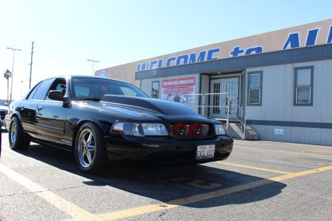 2003 Ford Crown Victoria - black full-size sedan with a custom red-accented grille and EVIL VIC plate is parked at an angle near a WELCOME to the racetrack building.