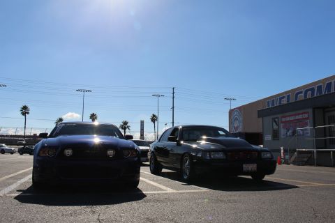 2003 Ford Crown Victoria - dark Ford Mustang and a black sedan are parked facing the camera under bright sun, with a WELCOME building to the right.