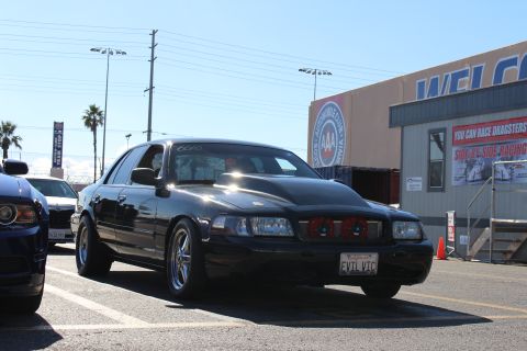 2003 Ford Crown Victoria - black full-size sedan with a custom grille and EVIL VIC plate is parked beside a blue car near a building with racing signage.