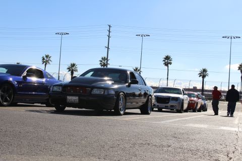 2003 Ford Crown Victoria - black sedan with red taillights sits beside a blue Mustang and silver Chrysler 300 in a lot as two people stand at the right.