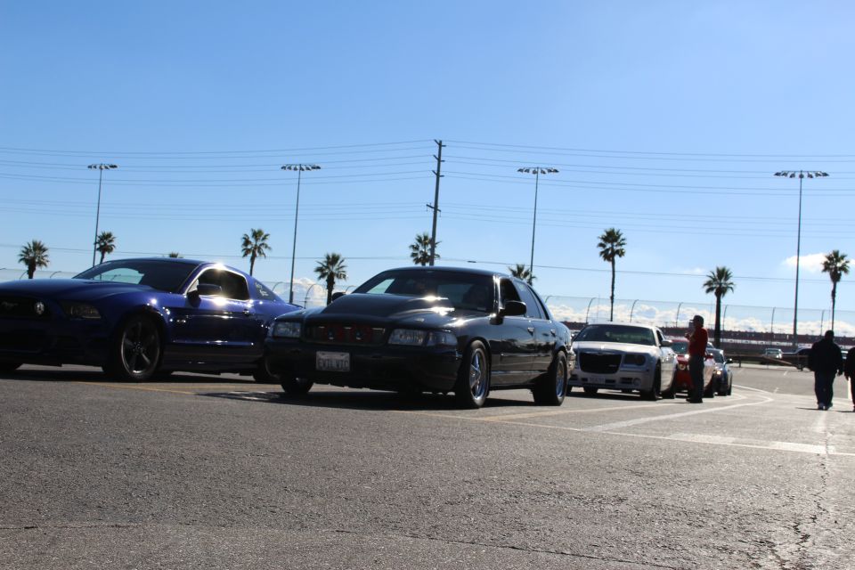 Blue Mustang and black sedan parked beside a silver Chrysler 300 with people in the lot and palm trees