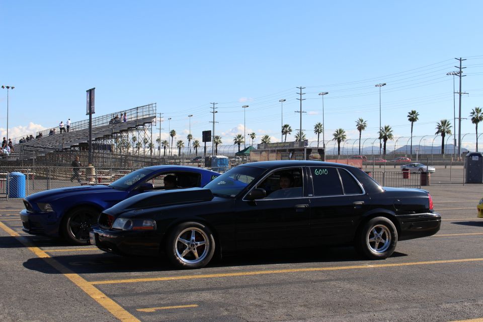 Black full-size sedan and blue Mustang in profile on a racetrack lot in front of empty grandstands