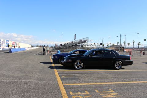 2003 Ford Crown Victoria - black sedan and a blue car sit along a wide drag strip lane in front of grandstands as a person stands at the trackside.