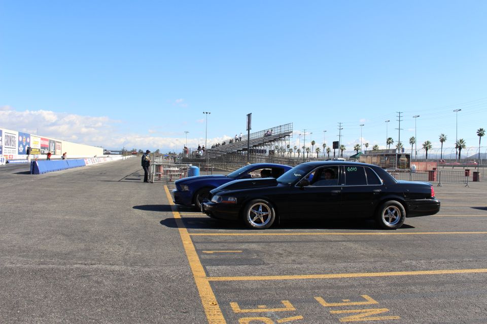 Black sedan and blue car parked along a drag strip lane with grandstands and a person standing nearby