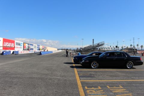 2003 Ford Crown Victoria - black full-size sedan is parked on a drag strip next to a wall with Lucas Oil and Auto Club Dragway signage and grandstands.