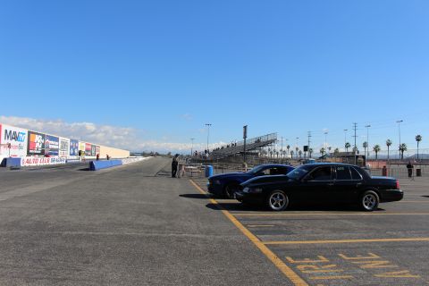 2003 Ford Crown Victoria - black sedan and a blue car sit at the edge of an empty drag strip with grandstands and trackside signage in the distance.