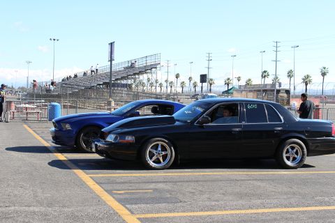 2003 Ford Crown Victoria - black full-size sedan marked 6010 on its window and a blue Mustang are shown in profile in a track parking area near grandstands.
