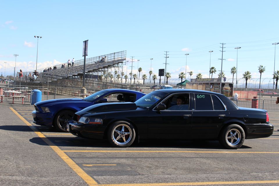 Black sedan numbered 6010 with driver inside, blue Mustang behind, in front of grandstands